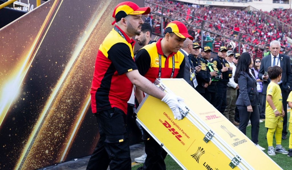Personal de DHL carga equipo en una ceremonia deportiva con presencia policial y público en el fondo.