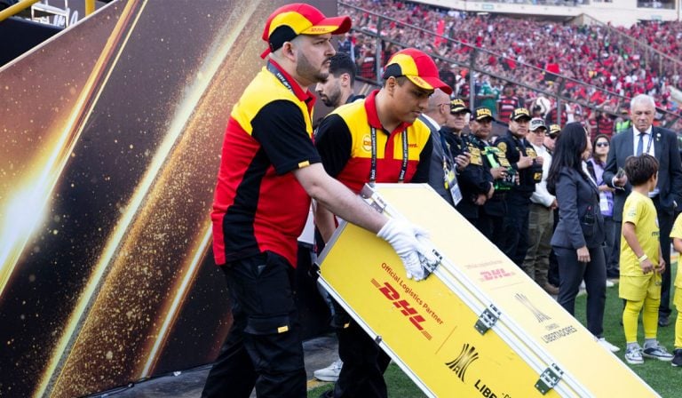 Personal de DHL carga equipo en una ceremonia deportiva con presencia policial y público en el fondo.
